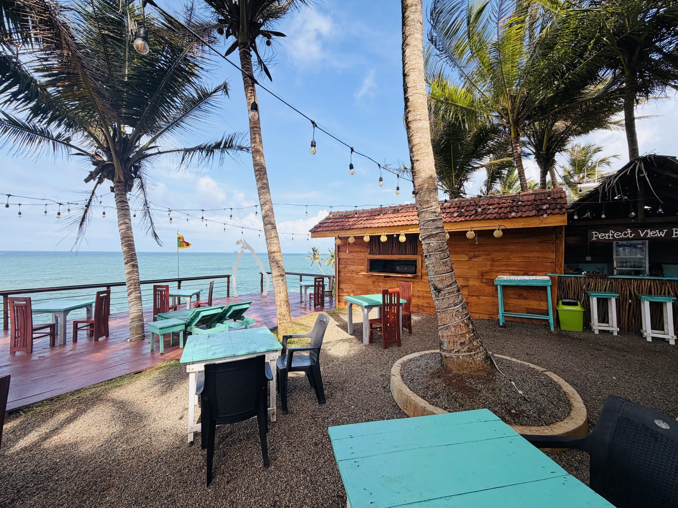 The bar hut and turquoise tables under the palms with the sea behind, on a bright day
